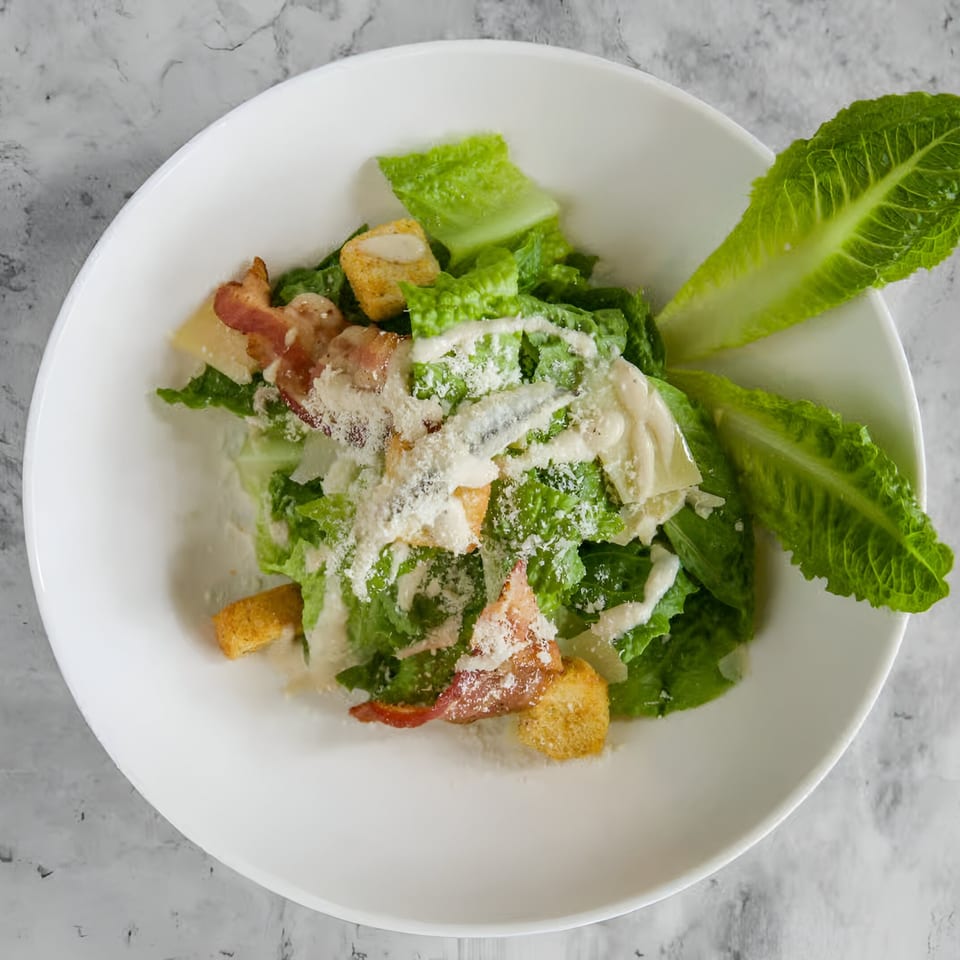 Caesar salad with romaine lettuce, bacon, croutons, parmesan, and dressing in a white bowl.