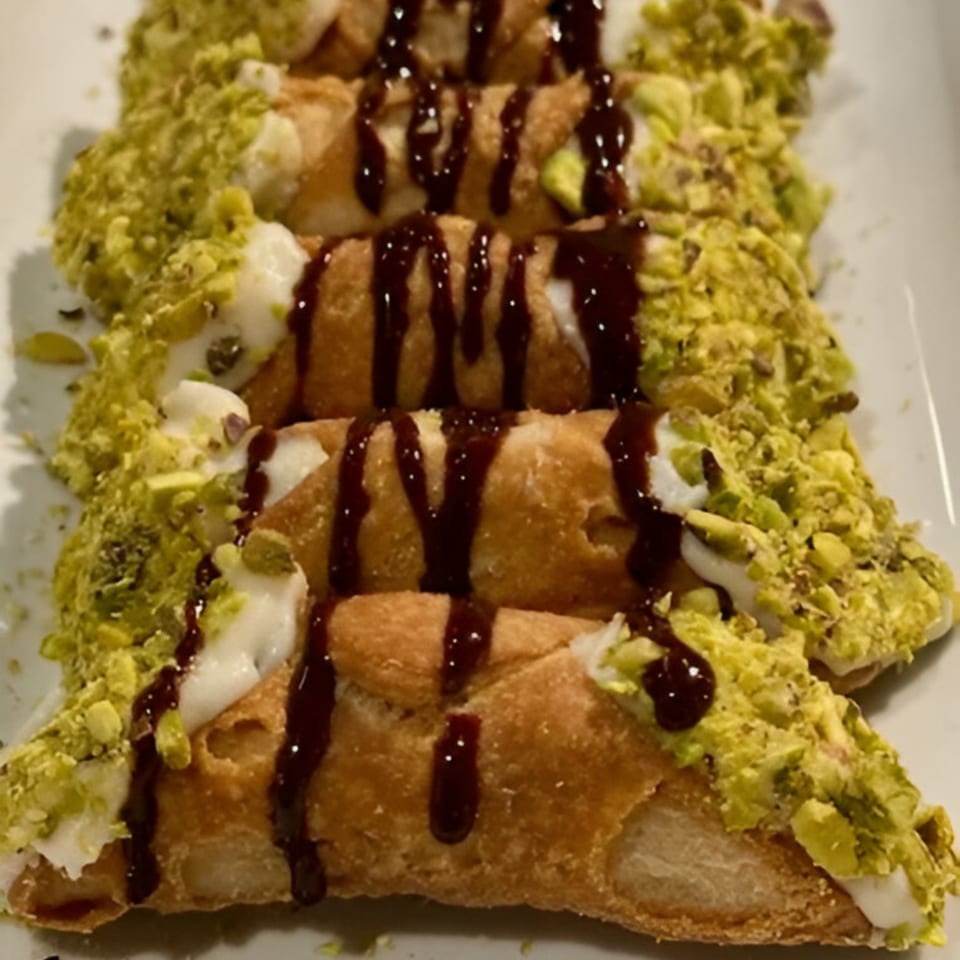 Five cannolis filled with white cream, drizzled with chocolate, and topped with crushed pistachios.